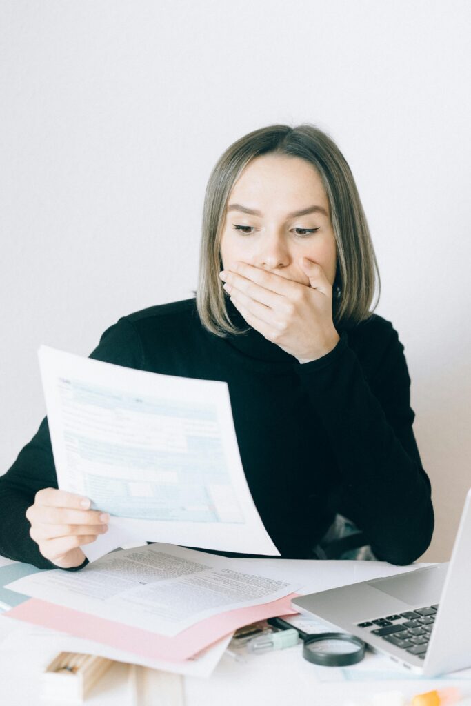 A shocked woman examines financial documents at her desk with a laptop indoors.