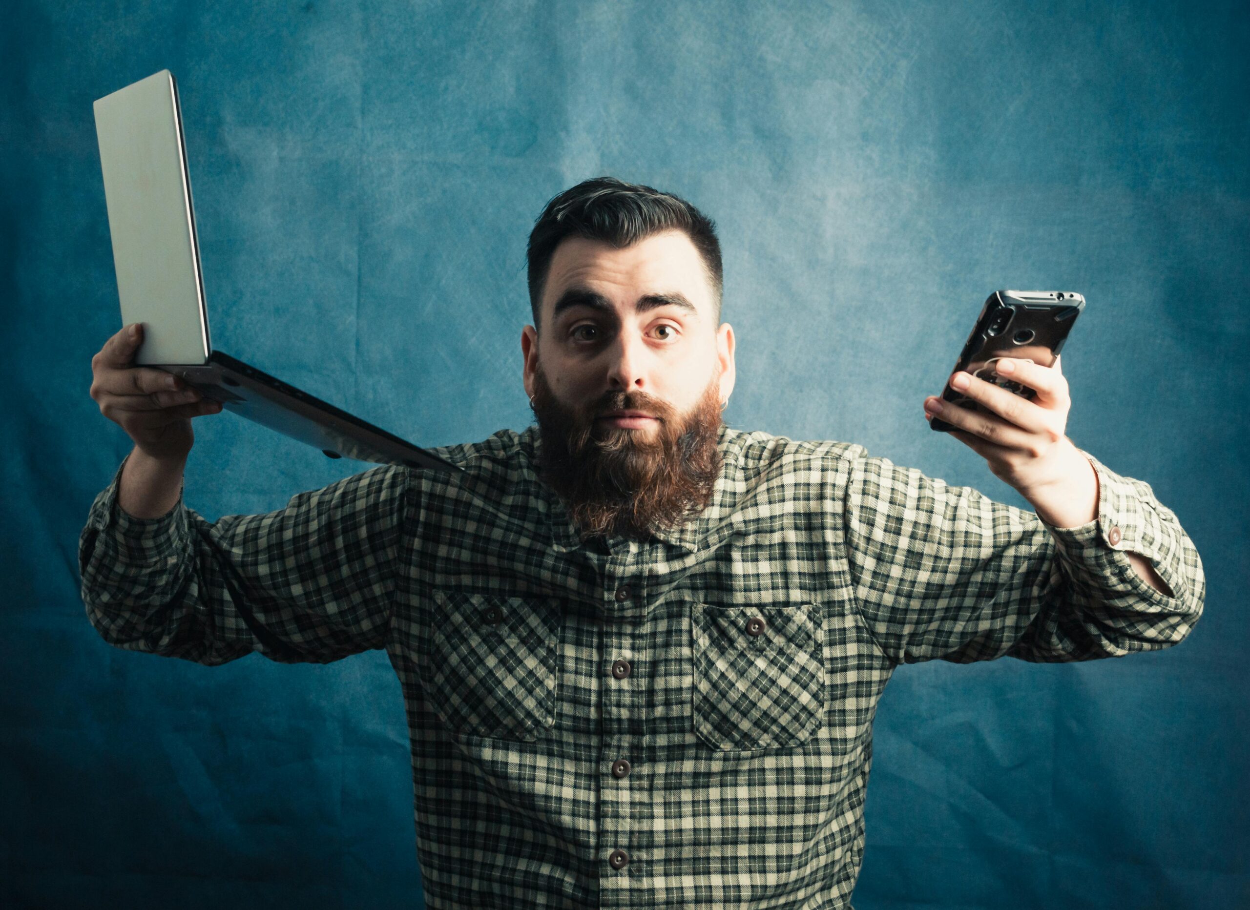 Man with a beard holding a laptop and smartphone against a blue background.
