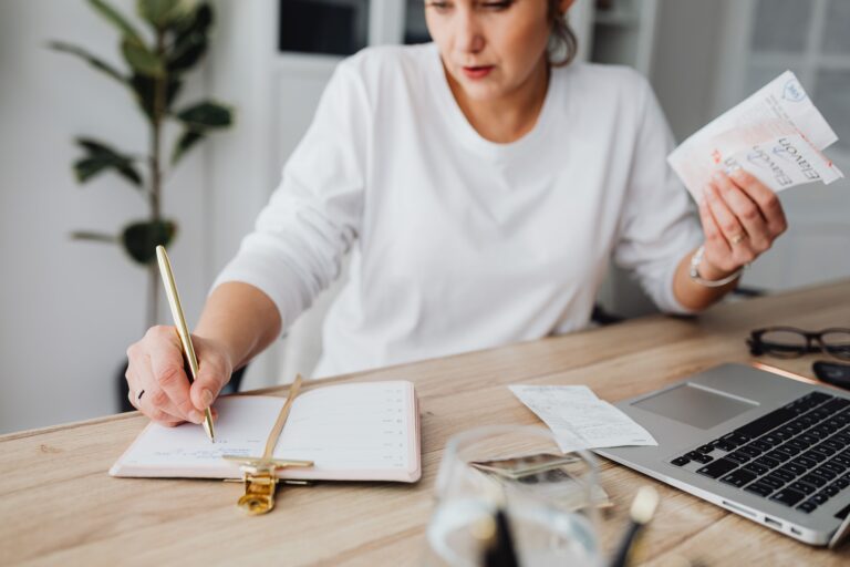 Woman managing finances, writing on notepad, and holding receipts at a desk.