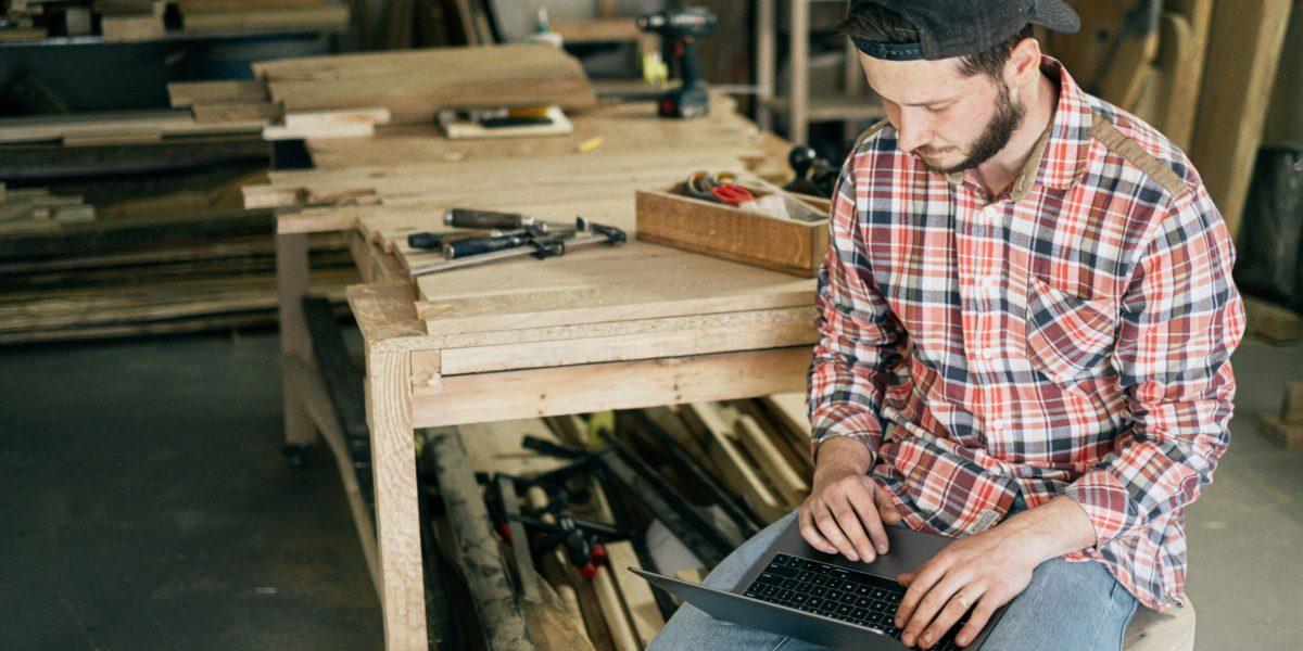 A carpenter working on his laptop in a wood workshop, surrounded by tools and materials.